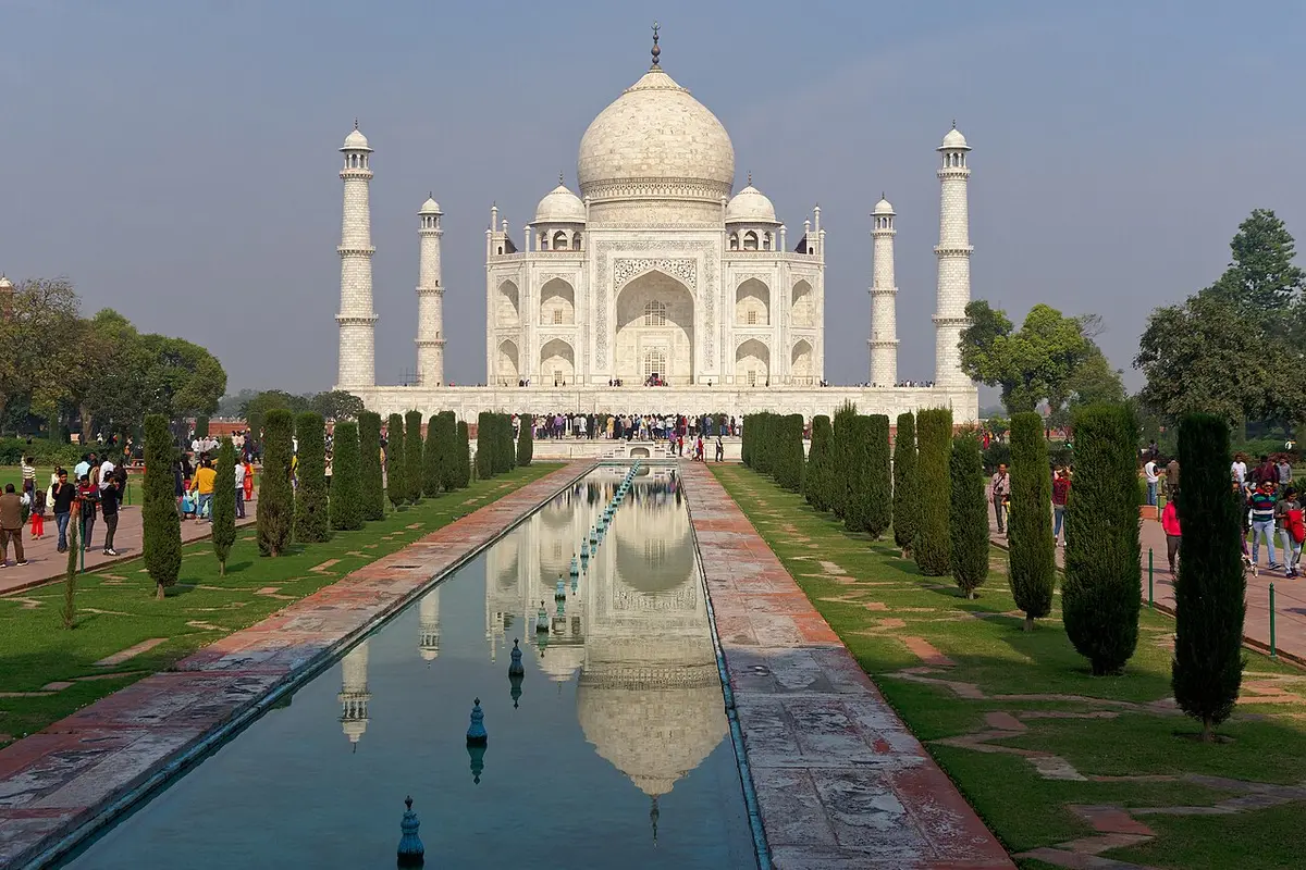 The Taj Mahal seen along its long reflecting pool, with cypress trees on both sides and visitors on the garden paths